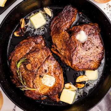 overhead view of seared steaks in cast iron skillet with butter, garlic and herbs