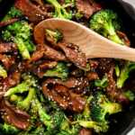 overhead view of wooden spoon scooping up beef and broccoli garnished with white sesame seeds