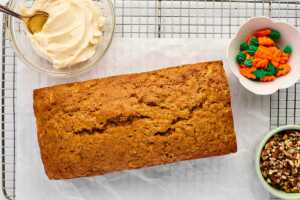 overhead view of baked carrot cake loaf on cooling rack with frosting, frosting carrots and chopped pecans in bowls
