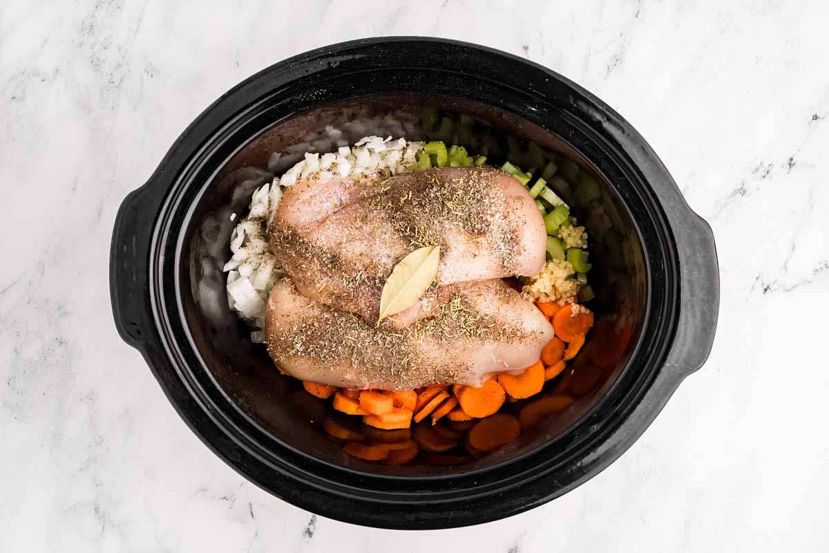 overhead view of raw vegetables and chicken breast in black slow cooker insert