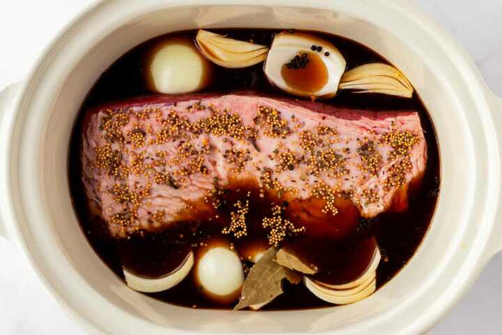 overhead view of uncooked corned beef brisket in slow cooker with broth and onions